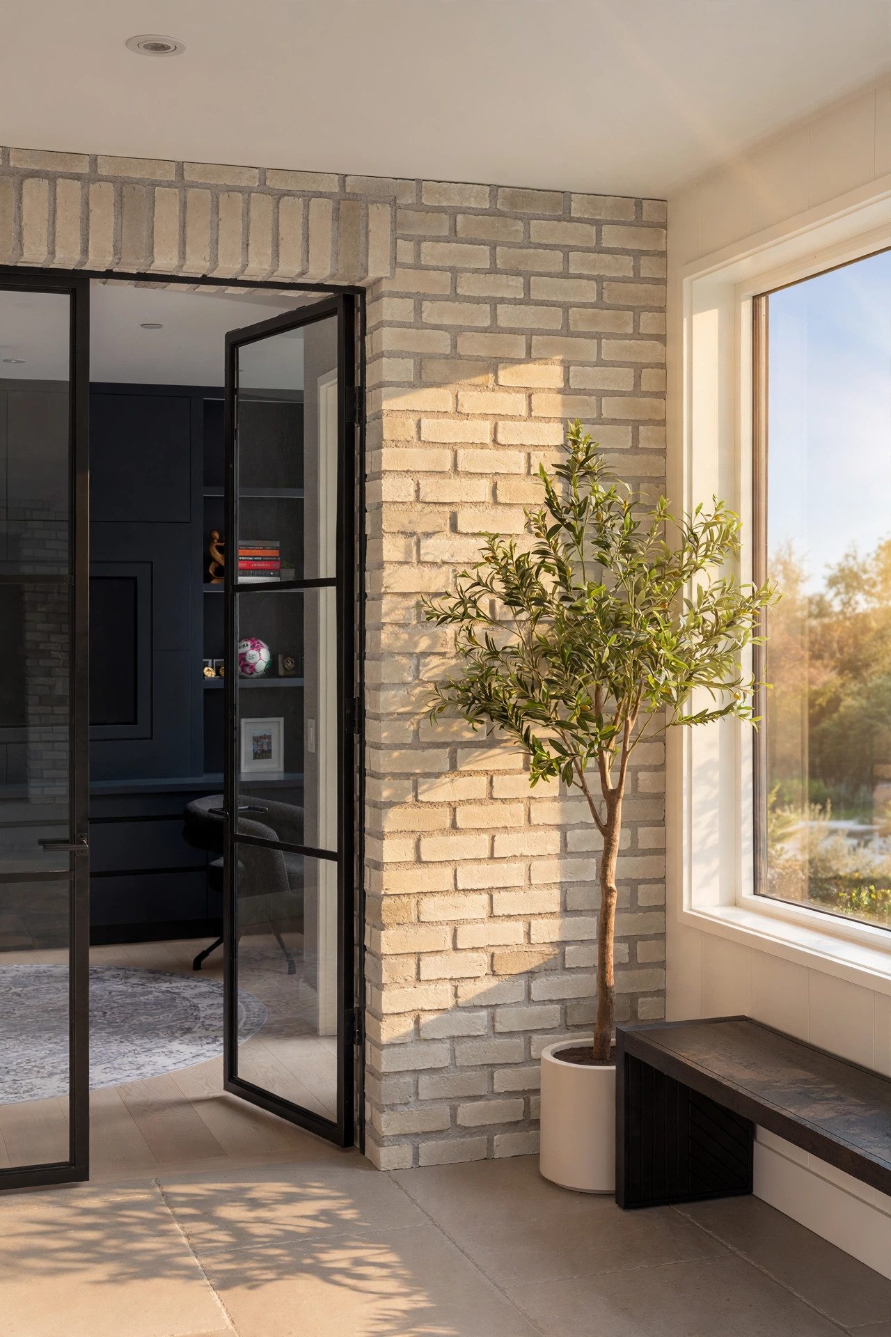 Sunlit white-brick passage with black steel doors and an indoor olive tree